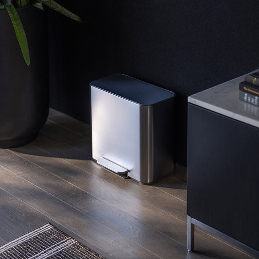 Simplehuman 16L Brushed Stainless Steel Mini Step Can with foot pedal, positioned on wood flooring next to a black storage cabinet with books next to it and plants on the other side.
