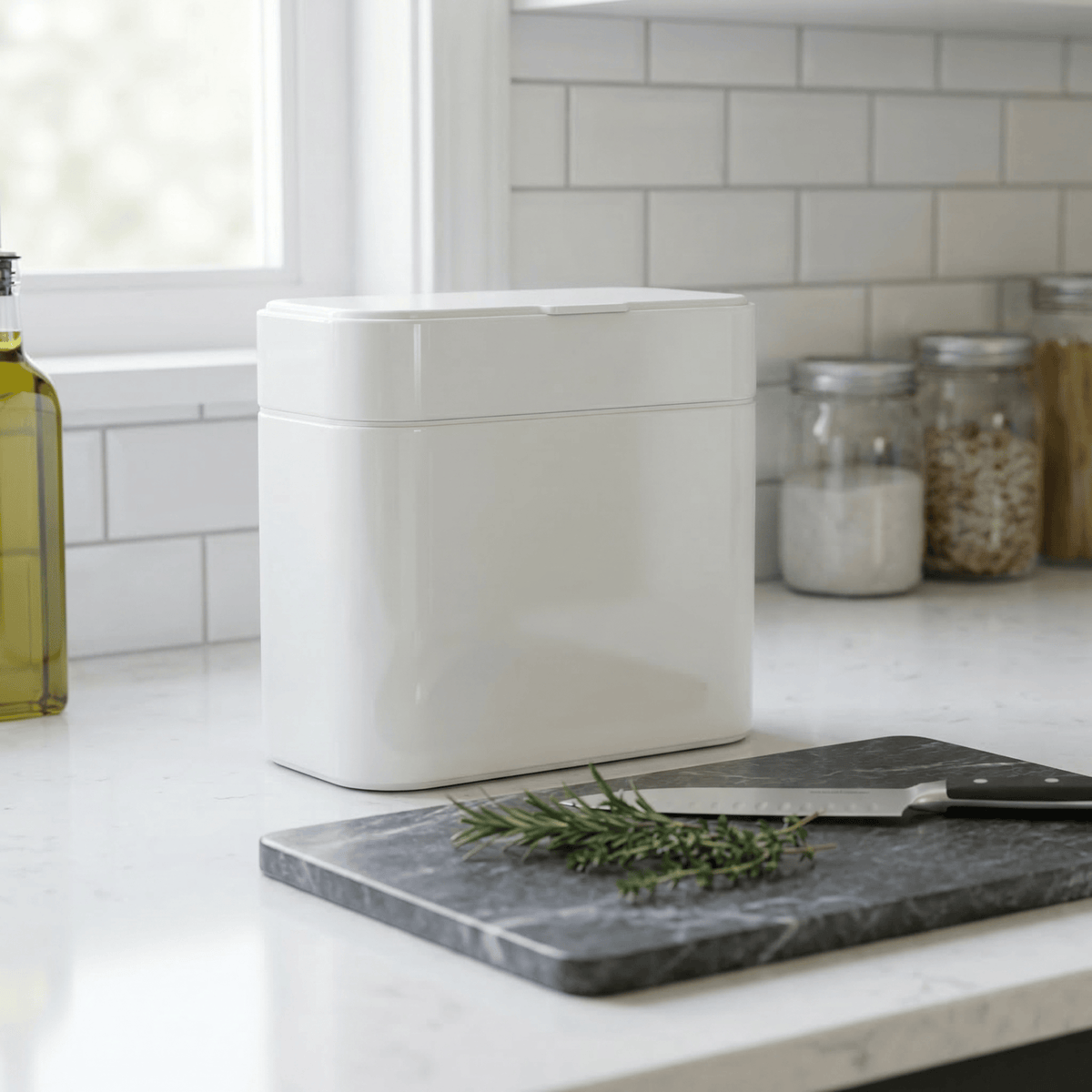 Simplehuman's White Compost Caddy in a bright, simple kitchen countertop settings around a bottle of oil and multiple mason jars.